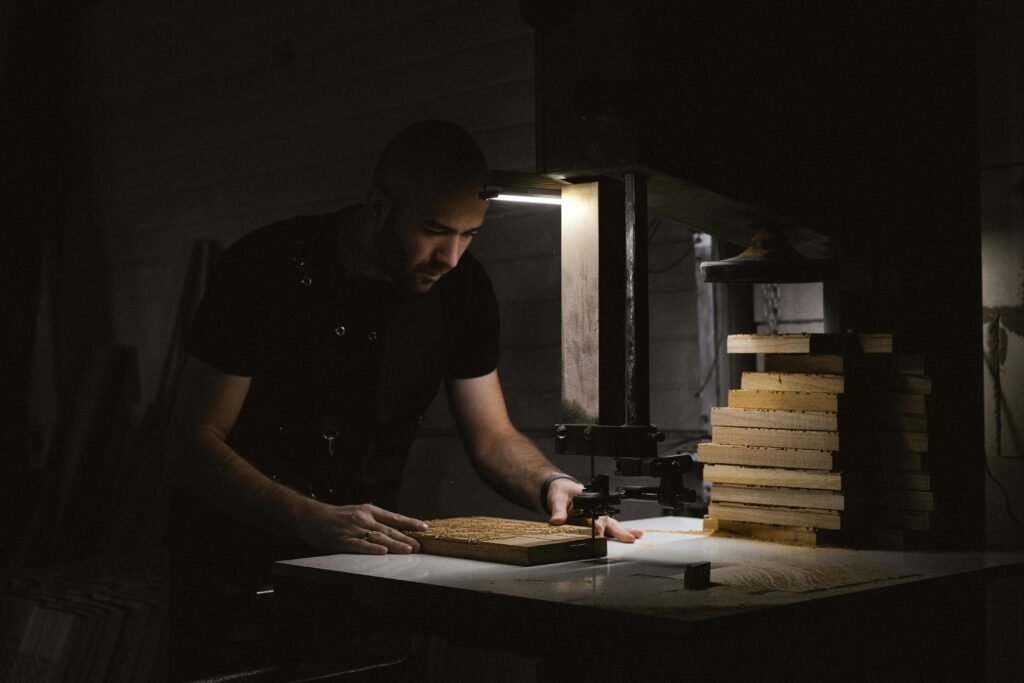 A skilled woodworker, concentrating while using a bandsaw in a dimly lit workshop with stacked wooden planks.